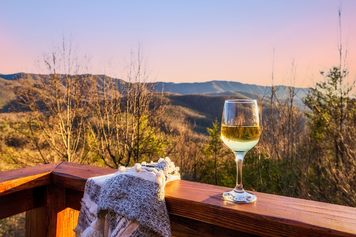 A glass of white wine rests on a wooden railing, accompanied by a textured blanket. The background features a breathtaking view of distant mountains under a soft evening sky, highlighting the natural beauty of the surrounding landscape.