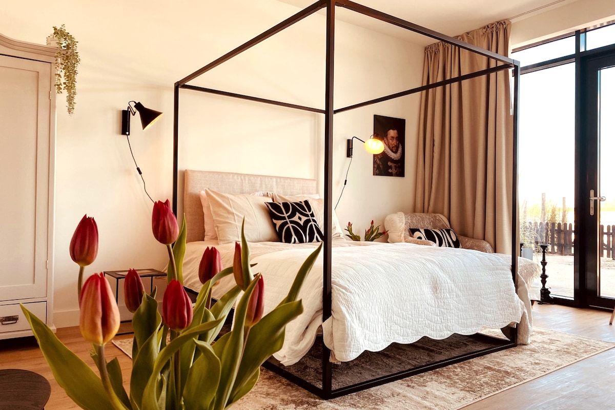 A modern bedroom features a four-poster bed adorned with a crisp white quilt, complemented by patterned pillows. Soft lighting is provided by wall-mounted lamps. A chair is positioned nearby, with fresh tulips adding a vibrant touch in the foreground. Large glass doors open to outdoor views.