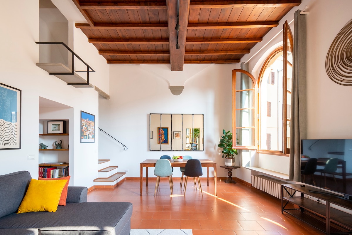 The bright living area features high wooden ceilings and large arched windows, allowing natural light to illuminate the space. A comfortable gray sofa is positioned near a sleek wooden dining table set for four. The room includes a Smart TV and decorative wall art, enhancing the inviting ambiance.