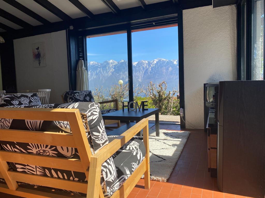 A bright living area features a wooden sofa with floral patterned cushions, facing large glass doors that provide an unobstructed view of distant mountains. A coffee table rests on a neutral rug, with a television unit visible in the corner.