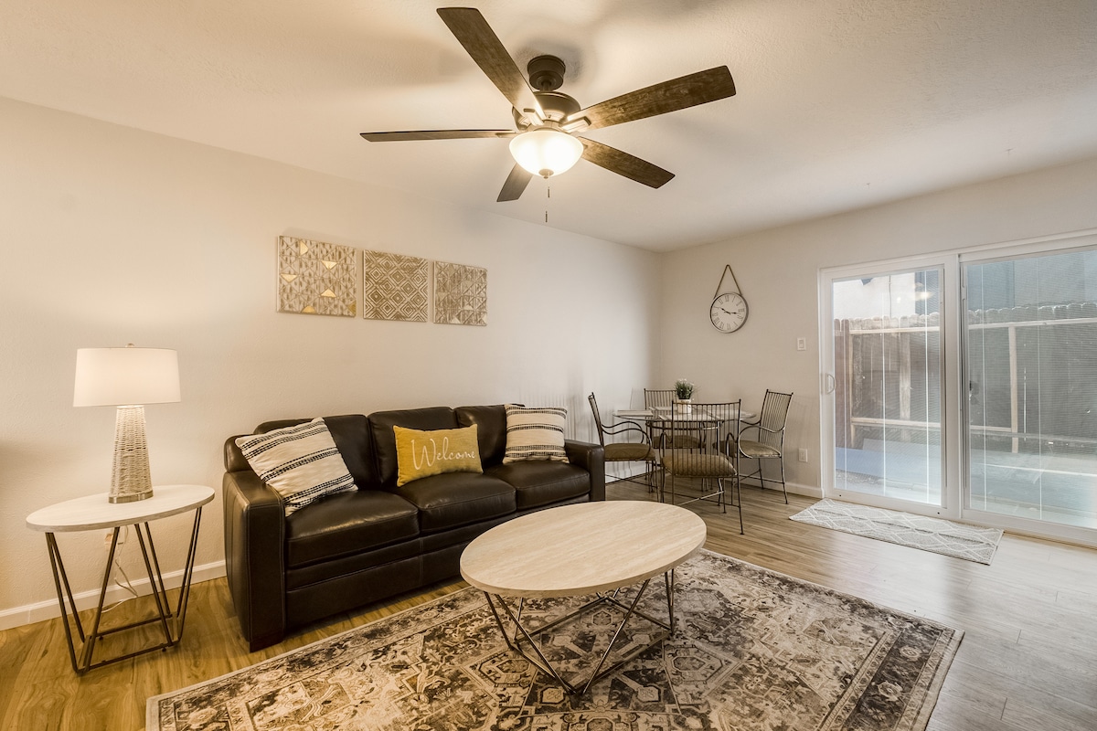 A clean and modern living space features a comfortable leather sofa and a round coffee table atop an area rug. A ceiling fan gently rotates overhead, while a dining area with a metal table and chairs is visible in the background. Large sliding doors lead to an outdoor area.