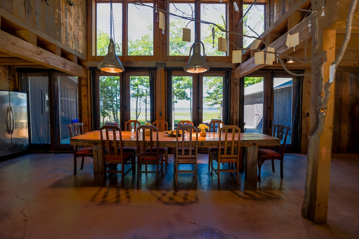 A spacious dining area features a long wooden table surrounded by wooden chairs, illuminated by pendant lights. Large windows showcase views of the outdoors, allowing natural light to flood the space. The floor is finished with a polished concrete texture.