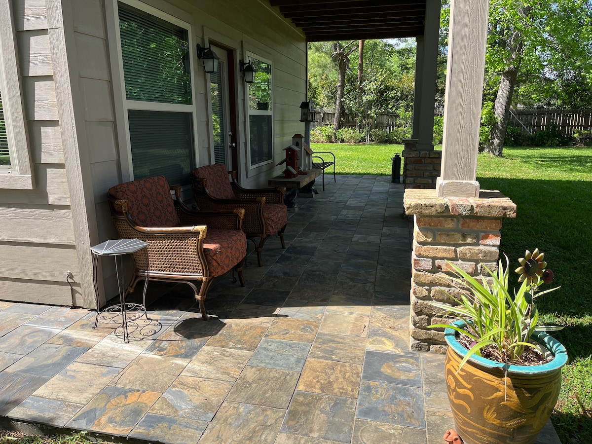 A covered patio area showcases two comfortable chairs with cushions, positioned against a backdrop of green foliage. A small side table rests beside the chairs, while a potted plant adds a touch of color to the space. The stone floor features varied shades of gray and brown.