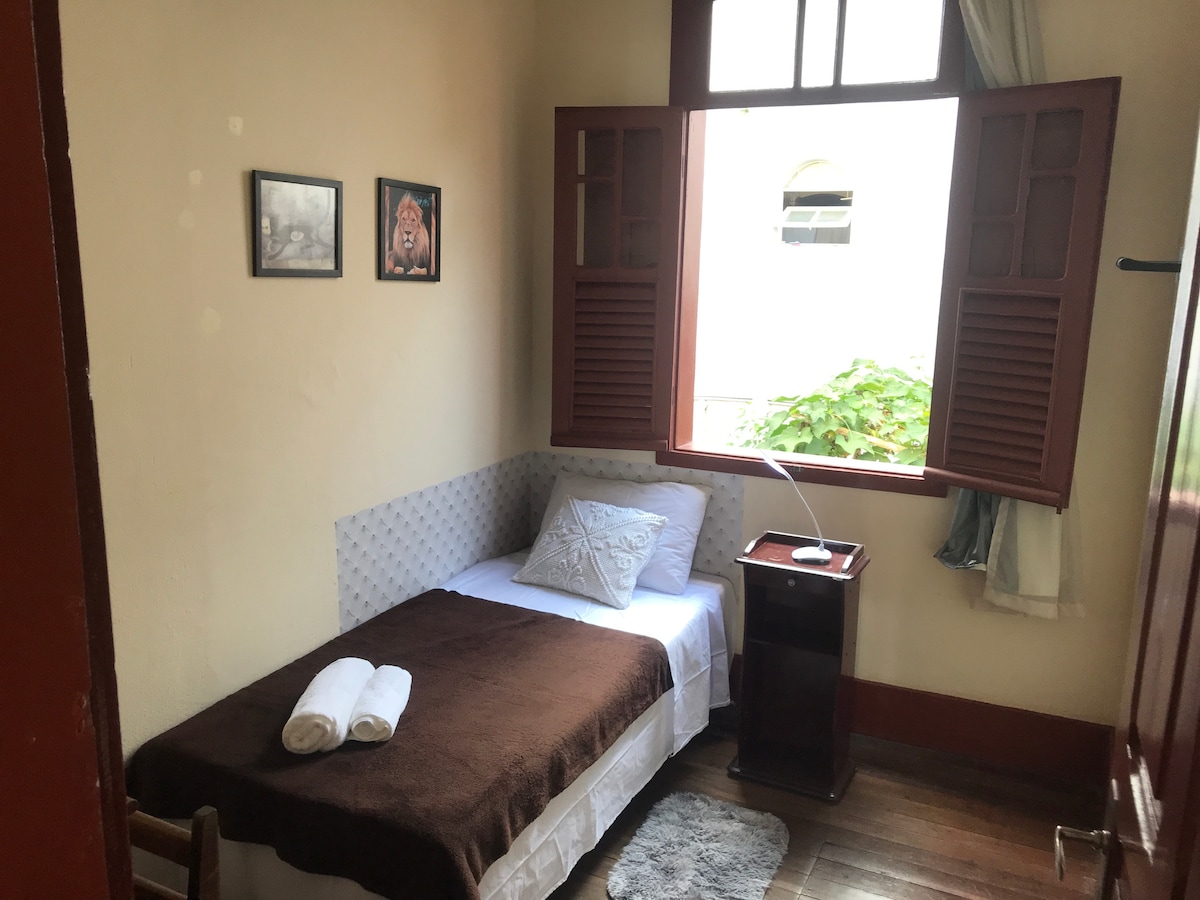 A cozy sleeping area is presented, featuring a single bed adorned with a brown blanket. A small bedside table holds a lamp, and fresh towels are neatly arranged. Natural light enters through the wooden window, highlighting the room's warm tones and simple decor.