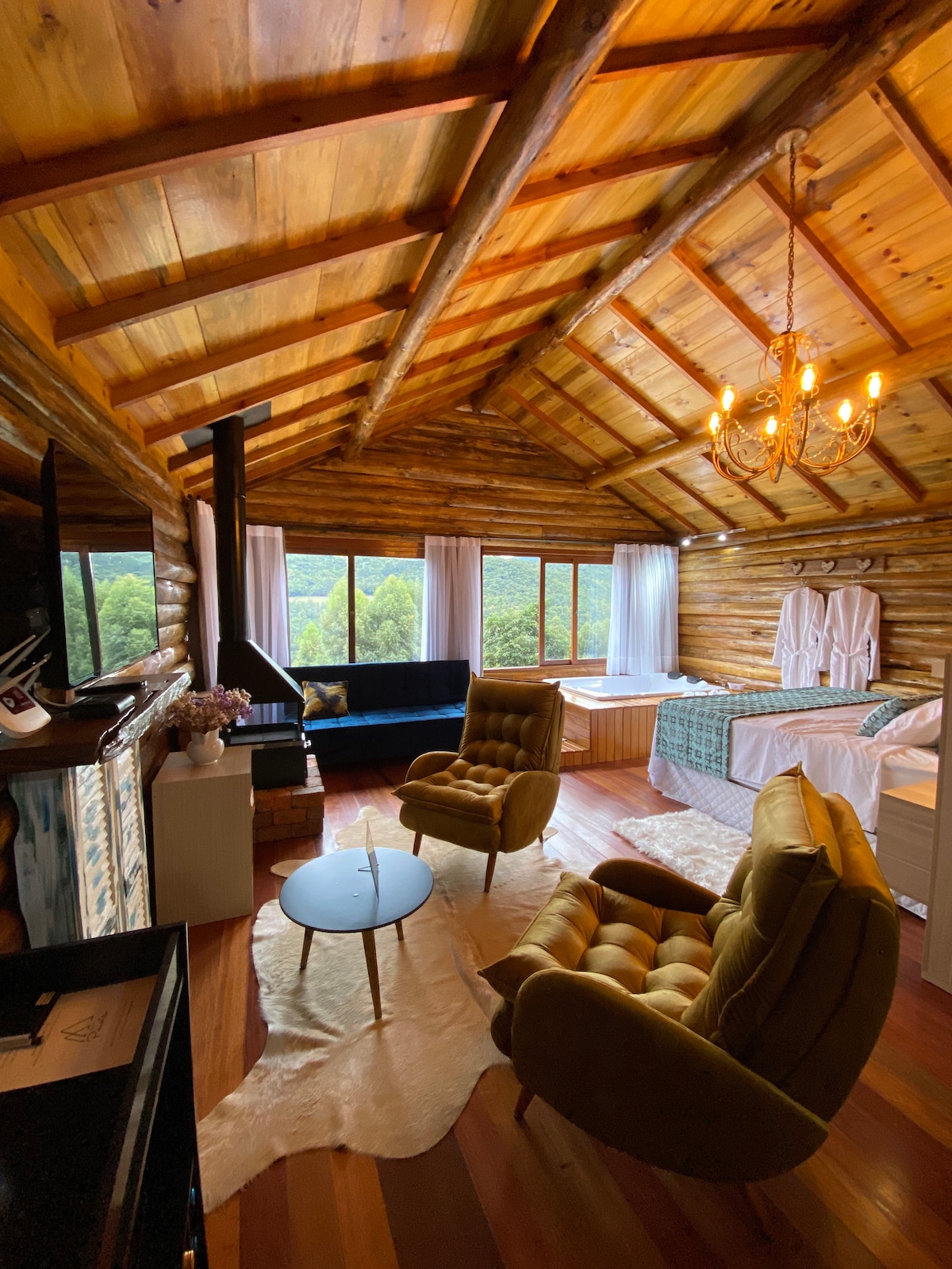 A rustic wooden cabin interior features a high ceiling with exposed beams. Two plush armchairs are placed in front of a modern fireplace. Large windows allow natural light to fill the space, offering views of the surrounding landscape. A cozy bed and a freestanding bathtub are also visible.