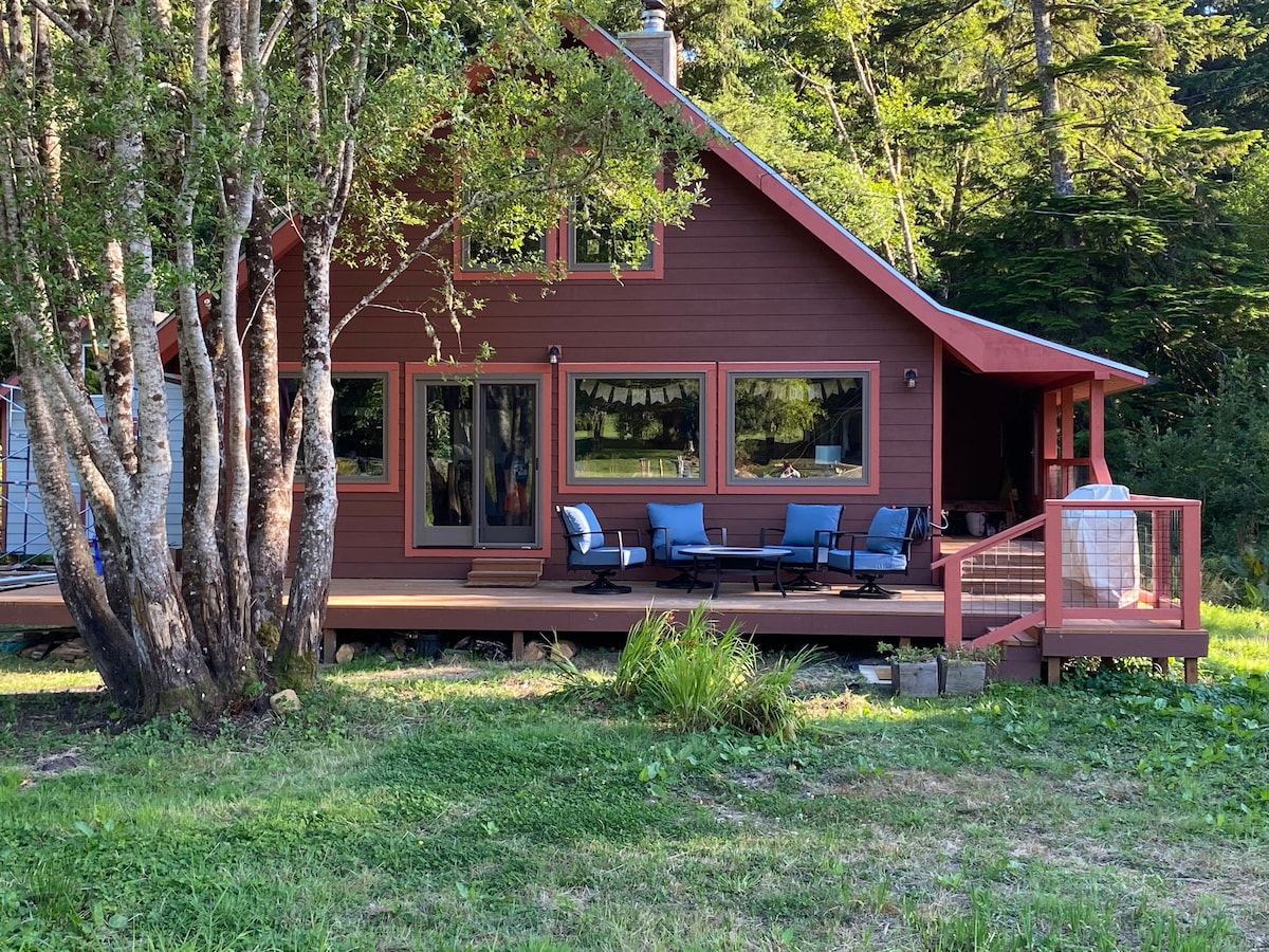 A charming red wooden house is set amidst greenery, featuring a spacious deck equipped with comfortable seating. Large windows invite natural light, while surrounding trees provide a peaceful, natural backdrop. The entrance is accessible from the deck, enhancing the connection to the outdoors.