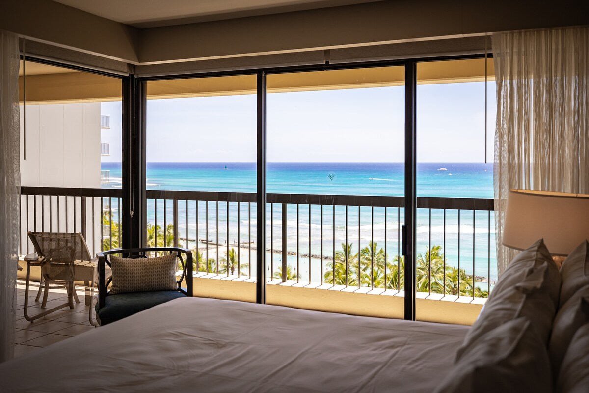 A spacious bedroom features large glass doors that open to a private lanai, offering expansive views of the ocean and surrounding palm trees. Natural light floods the room through sheer curtains, creating a tranquil ambiance, while a comfortable bed invites relaxation.