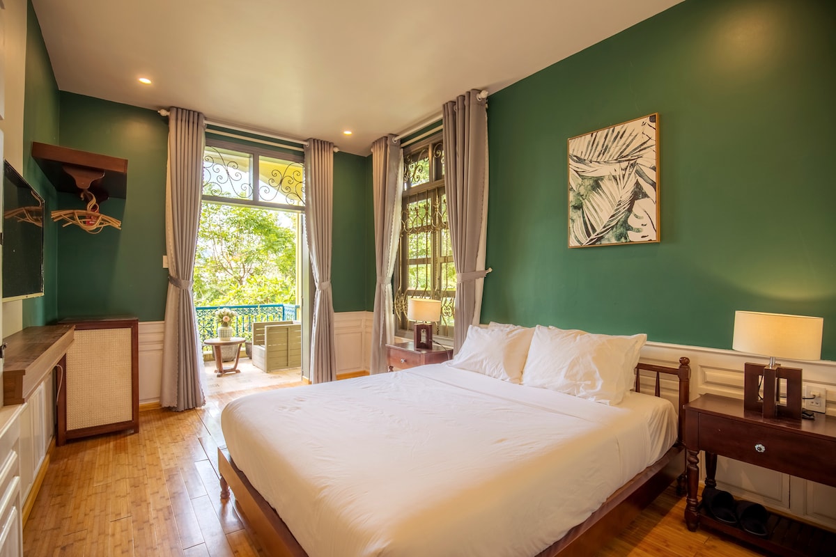 A spacious bedroom features a large bed with crisp white linens. Natural light pours through the balcony doors, revealing outdoor greenery. The walls are painted a calming green, complemented by wooden details. Two bedside tables with lamps flank the bed, enhancing the room's inviting ambiance.