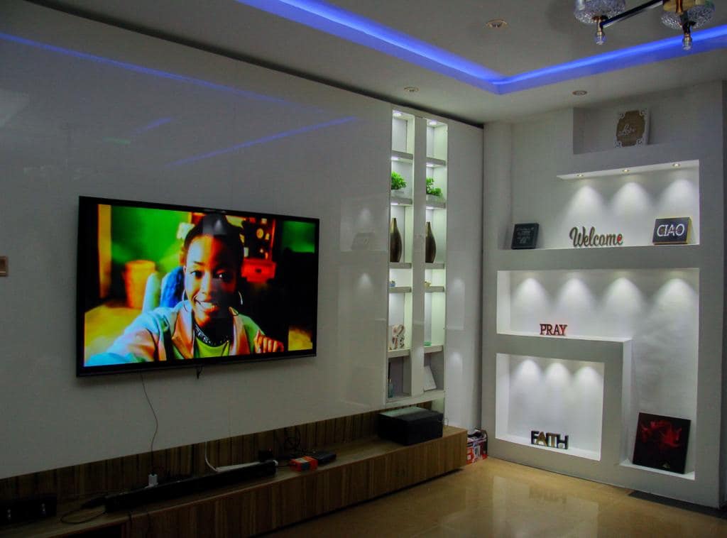 A modern living area features a large wall-mounted television, which displays vibrant colors. Sleek white shelving units hold decorative items, illuminated by soft lighting. The space is enhanced by elegant wall decor, creating a welcoming environment.