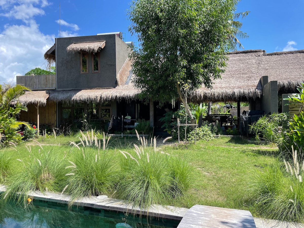 A tropical retreat is surrounded by lush greenery, featuring thatched roofs and natural materials. The structure includes wide outdoor spaces for relaxation, with a swimming pool visible in the foreground, harmonizing with the vibrant landscaping.