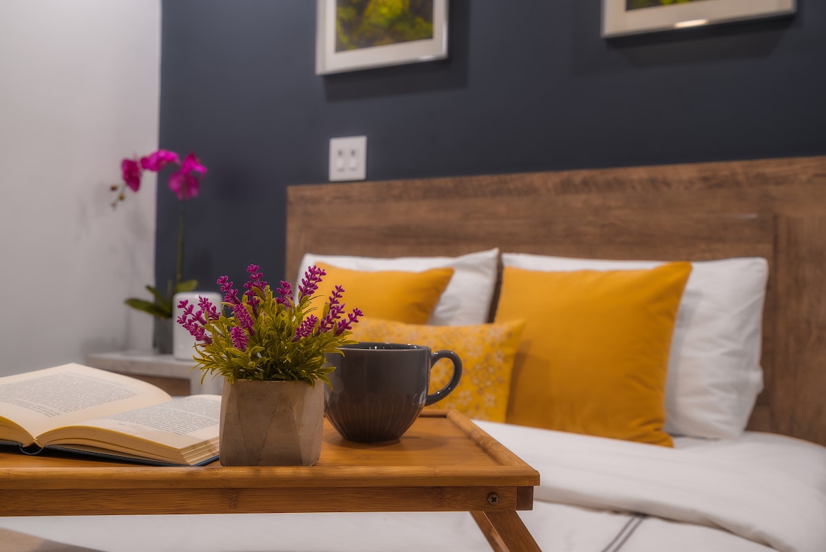 A cozy bedside table is presented, adorned with a small potted plant and a cup. An open book rests nearby, creating a tranquil reading space. The bed features bright yellow pillows against a dark wall, adding a touch of color to the room's design.