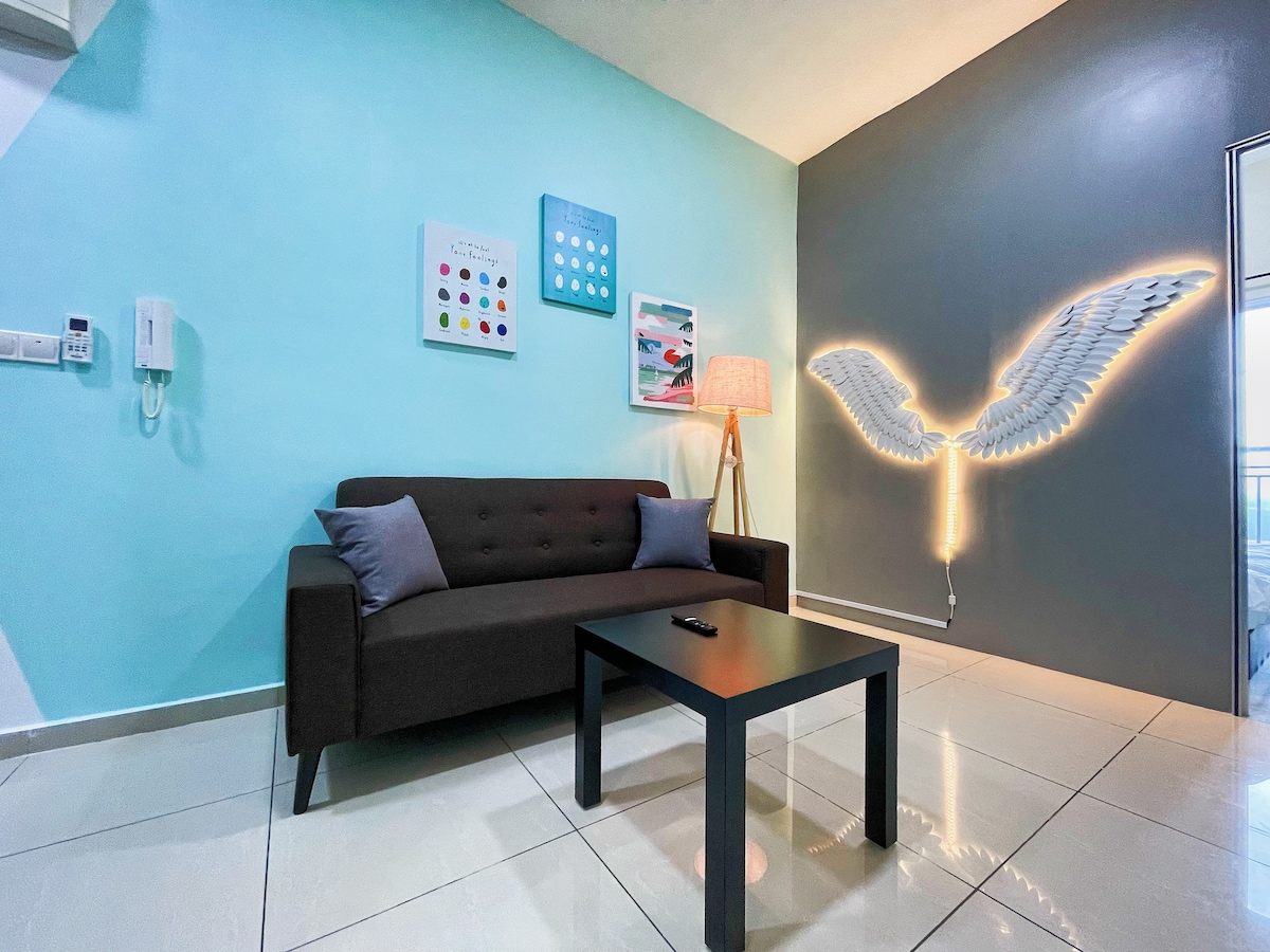 A modern living room features a dark sofa adorned with accent pillows, accompanied by a small black coffee table. A floor lamp adds warmth to the space, while colorful wall art and a lighted wing sculpture create visual interest against a two-tone blue and gray wall.