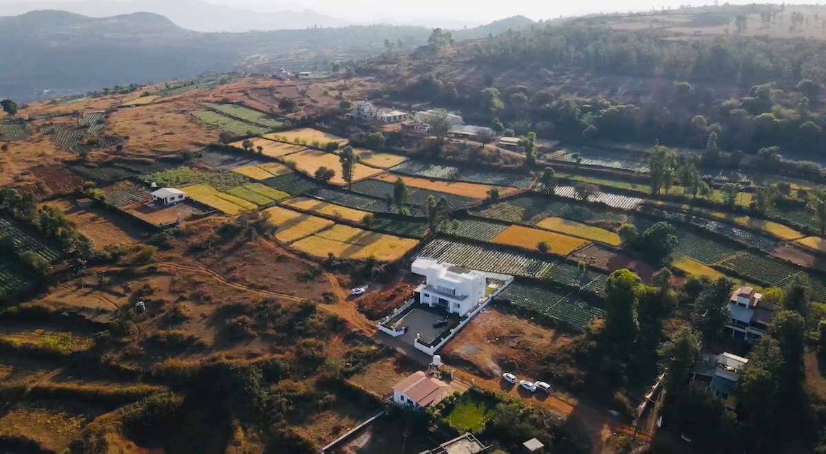 An aerial view captures a sprawling landscape of cultivated fields, featuring patchworks of green and golden crops. A modern white structure is positioned amidst the greenery, with distant hills visible under a bright sky, creating a serene rural setting.