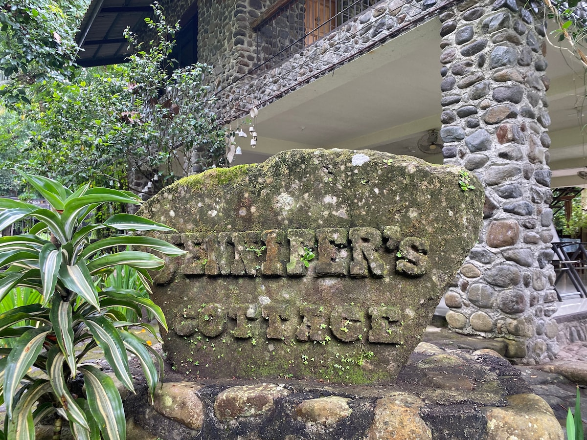A large stone sign displaying 'Jennifer's Cottage' is nestled among lush greenery. The textured surface is partially covered with moss, contributing to the natural ambiance. In the background, the stone facade of the cottage can be seen, adding to the rustic charm of the location.