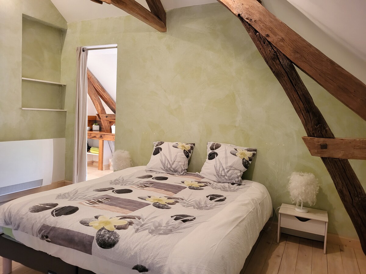 A cozy sleeping area features a spacious bed with a floral-patterned duvet. The green walls and natural wooden beams create a calming ambiance. A small nightstand with a lamp is placed beside the bed, while soft decorative pillows add texture to the overall decor.