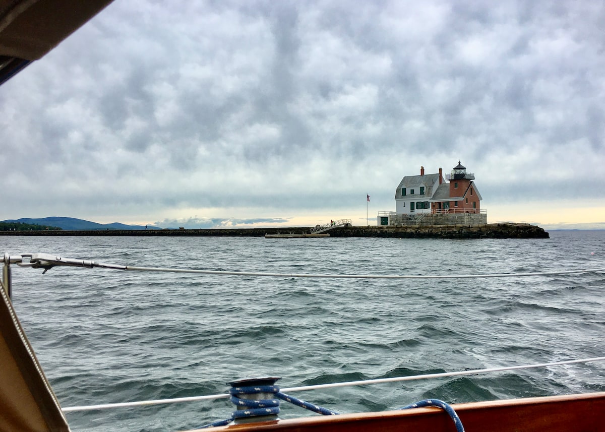 A historic house with a lighthouse stands prominently on a rocky shoreline, surrounded by gentle waves. The structure features a pink and white exterior with a tall, pointed roof, while a flag flutters in the breeze. A cloudy sky adds an atmospheric backdrop to the scene.