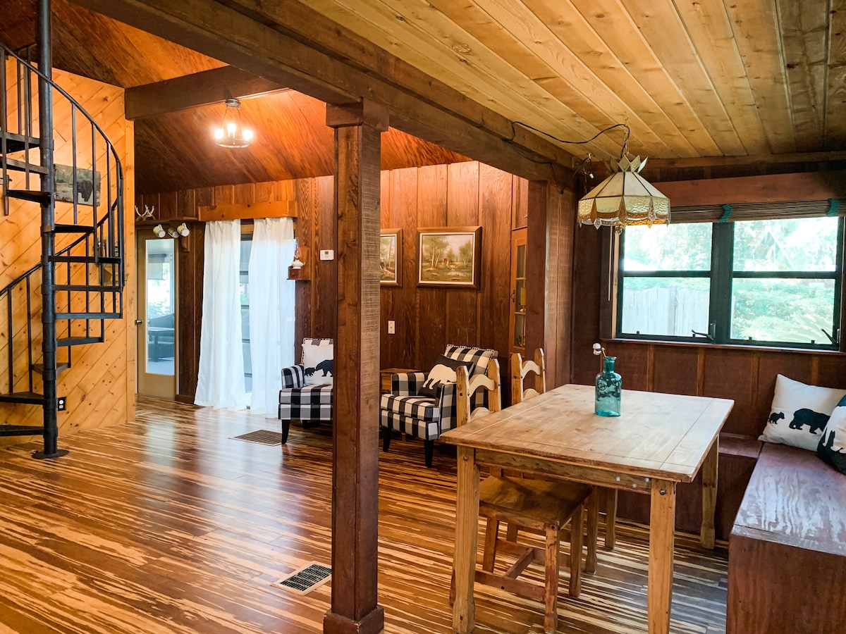 A rustic interior showcases wooden beams and warm-toned flooring, with a spiral staircase in the corner. A dining table is surrounded by chairs, while a cozy seating area with checkered upholstered chairs invites relaxation. Natural light filters through large windows adorned with sheer curtains.