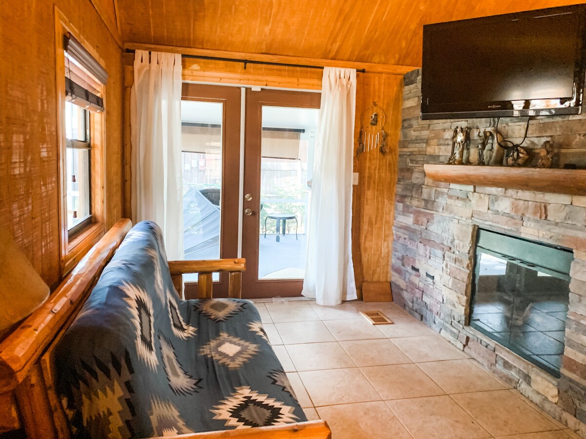 A cozy living area features a futon and a stone fireplace. Double doors lead to the porch, allowing natural light to brighten the space. A television is mounted on the wall, and light curtains frame the doors for a soft touch.