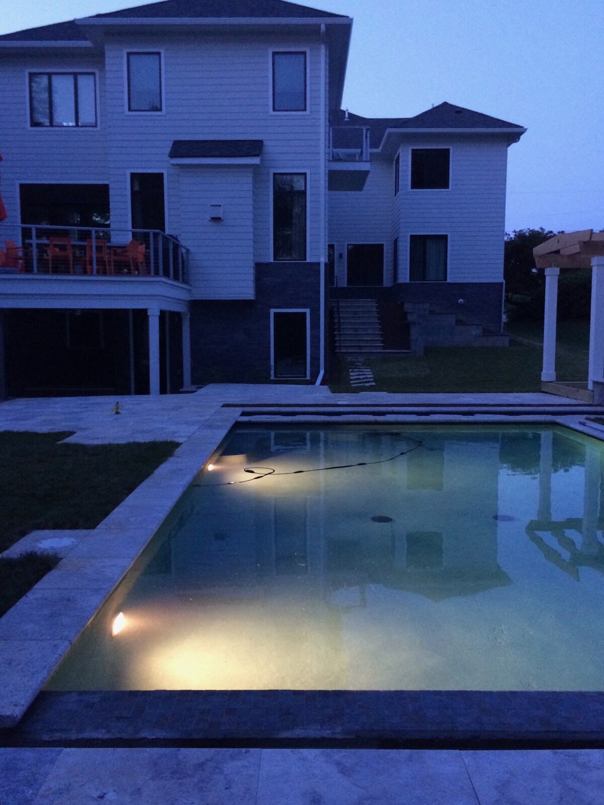 An elegant large home is depicted in the evening, with a well-lit pool reflecting the soft glow of underwater lights. The surrounding patio features a stone finish, and the exterior showcases multiple floors and expansive windows, creating a welcoming atmosphere.
