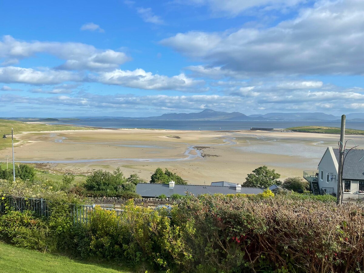 A panoramic view captures the expanse of Clew Bay, with sandy shores and distant hills under a partly cloudy sky. Lush greenery from the garden frames the scene, enhancing the natural landscape of the coastal area.