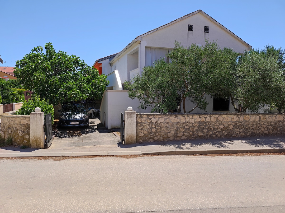 A two-story white building is visible, surrounded by lush greenery. A stone wall encloses the property, providing privacy. A shaded parking area can be seen in front of the building, featuring a parked car. The clear blue sky above adds a bright and open feel.