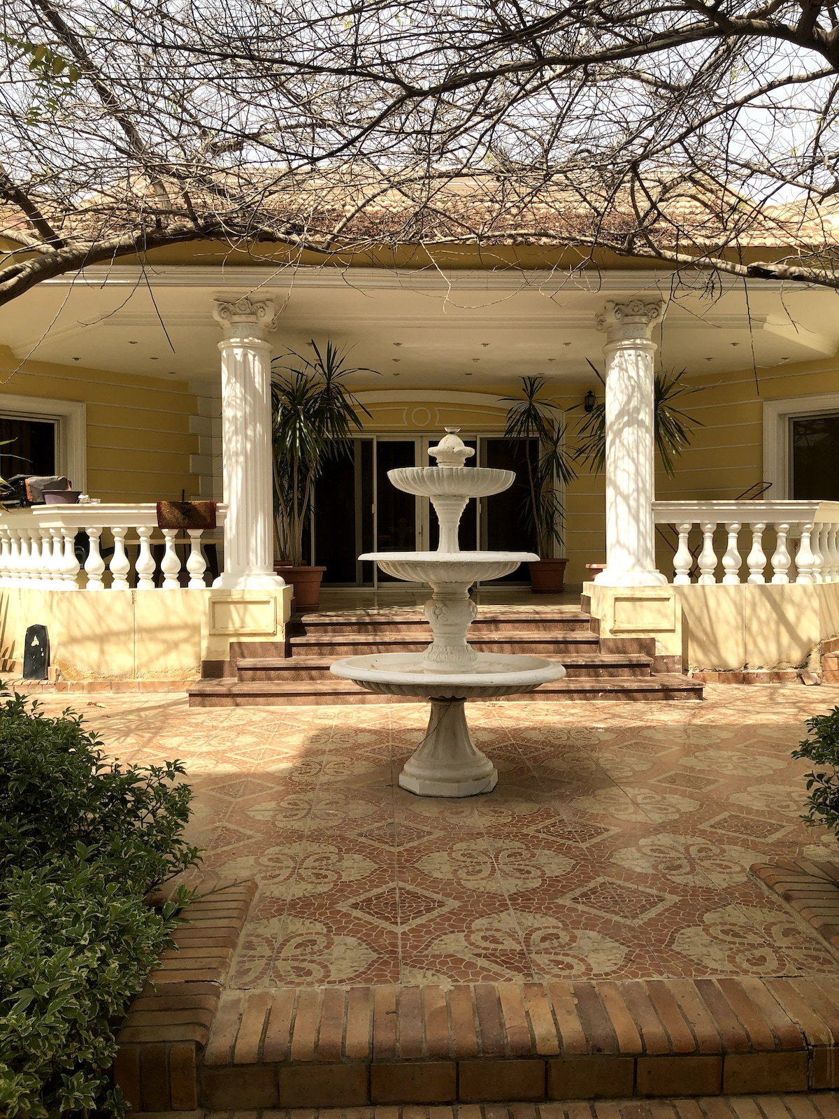 A charming entrance features a white stone fountain surrounded by lush greenery. The porch is supported by elegant columns, and large potted plants add to the welcoming ambiance. Decorative tiles pave the pathway leading to the main entrance.