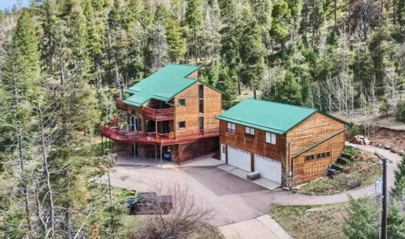 The exterior of the property displays a wooden two-story home with a green roof, complemented by a smaller building with a garage. Surrounded by tall evergreen trees, the driveway leads to the entrance, offering a peaceful mountain retreat ambiance.