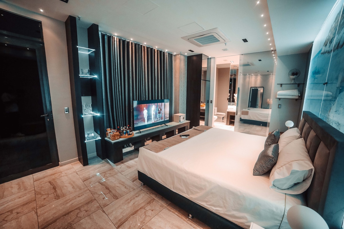 A modern bedroom features a spacious bed with subtle bedding, a large flat-screen television mounted on a dark wooden entertainment unit, and accents of soft lighting. The room is designed with marble flooring and mirrors that enhance the sense of openness.