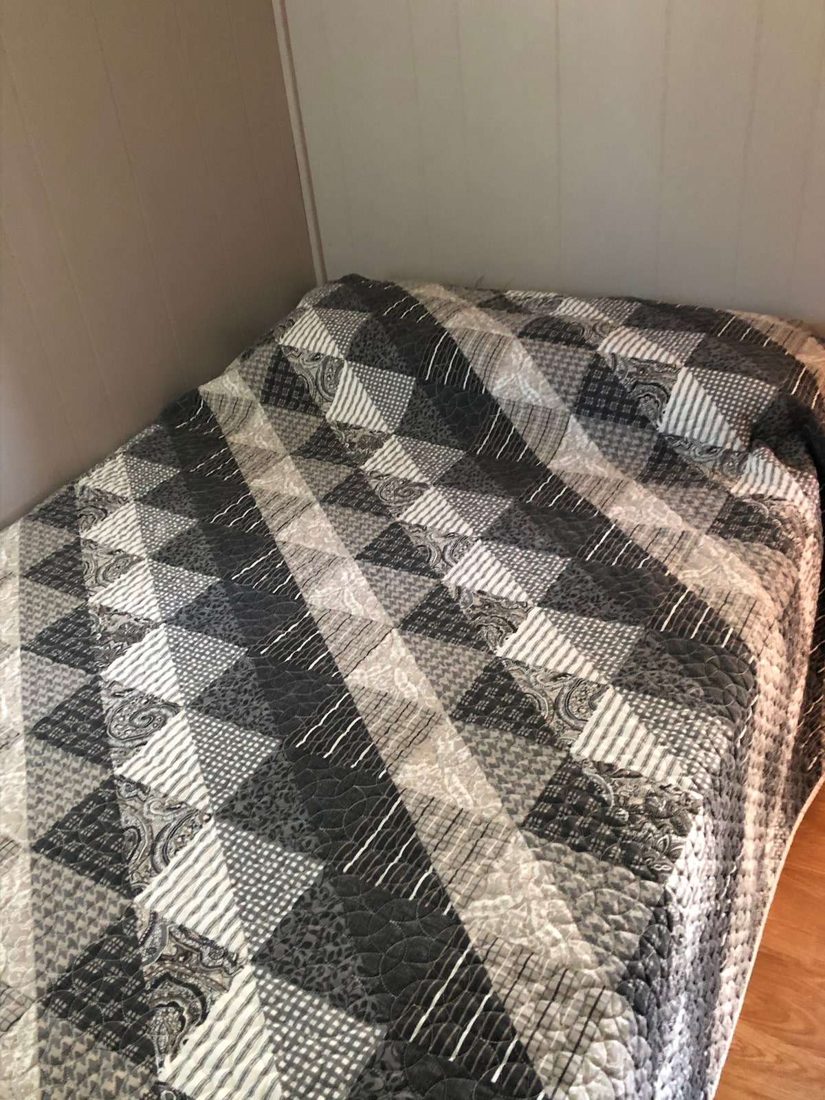 A cozy sleeping space features a double bed covered with a textured quilt in shades of gray and white. The simple wooden floor complements the neutral tones of the room, while walls are finished in light, natural hues.