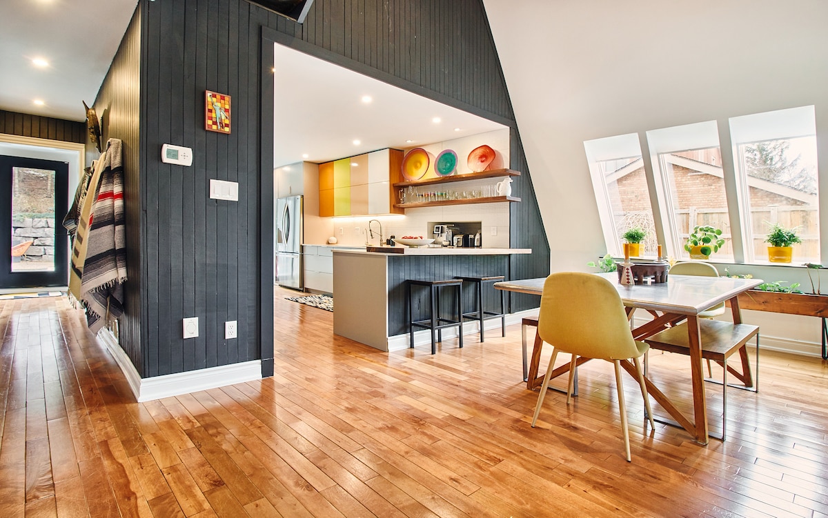 An open-plan kitchen and dining area features modern cabinetry with colorful dishware displayed on a shelf. Natural light floods through multiple windows, illuminating the warm wooden floors and contemporary furniture, including a dining table and chairs. A cozy, welcoming atmosphere is maintained throughout the space.
