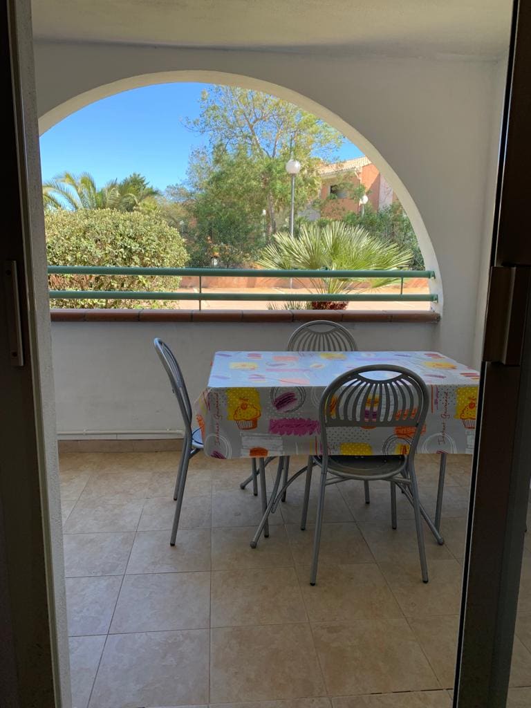 An arched terrace is visible, featuring a round table covered with a colorful tablecloth. Four silver chairs surround the table, and lush greenery is seen in the background, enhancing the serene outdoor space.