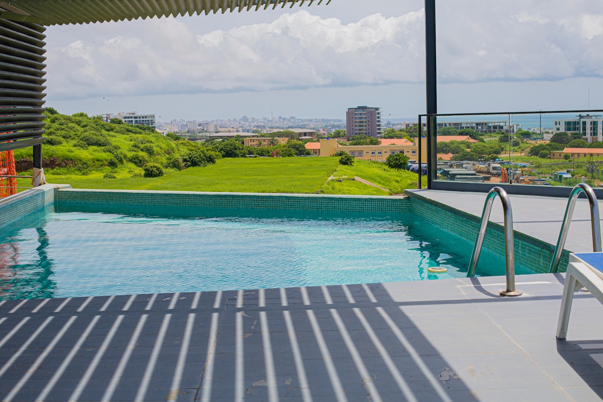 An unsupervised swimming pool is featured on the rooftop, bordered by sleek tiles. Sunlight casts gentle shadows from the overhead structure while panoramic views of the city and surrounding greenery are visible in the background, enhancing the open atmosphere.