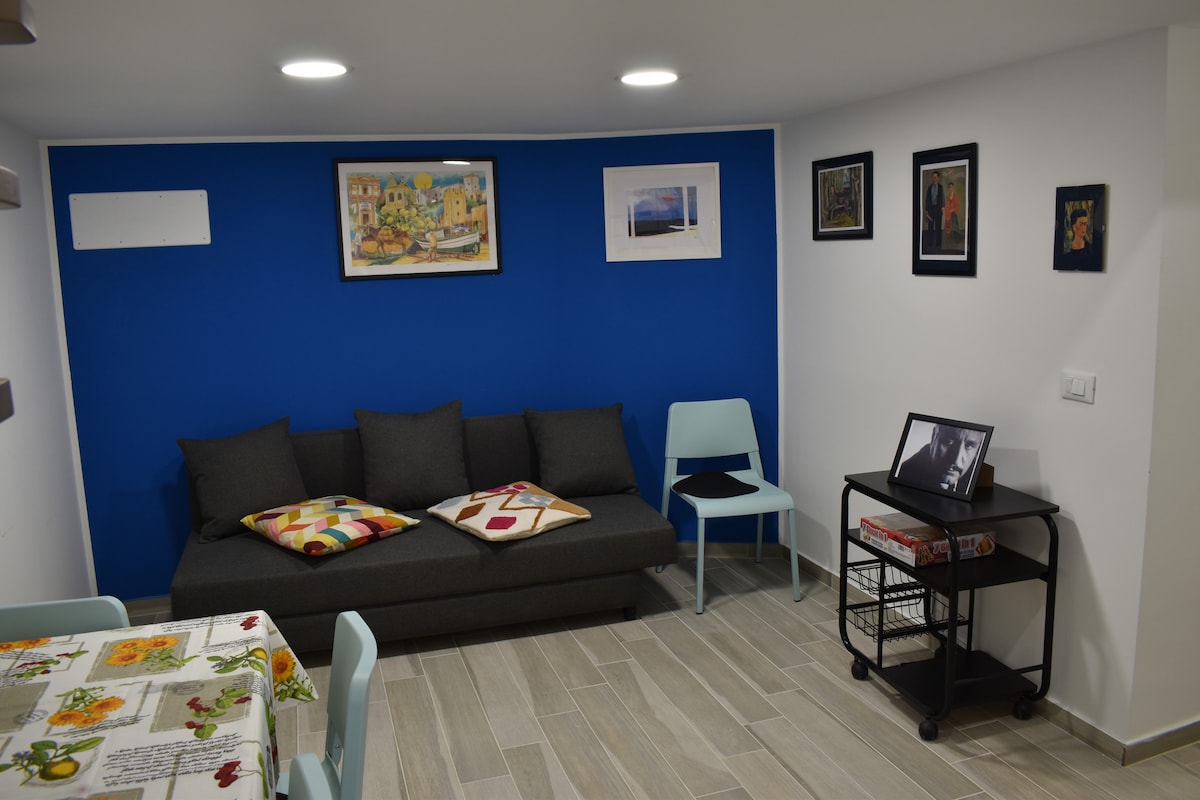 A modern living area features a dark gray sofa adorned with colorful cushions, complemented by a light blue wall. A small dining table is set nearby, while framed artwork decorates the walls, adding character to the space. A rolling cart holds additional items, providing convenience.