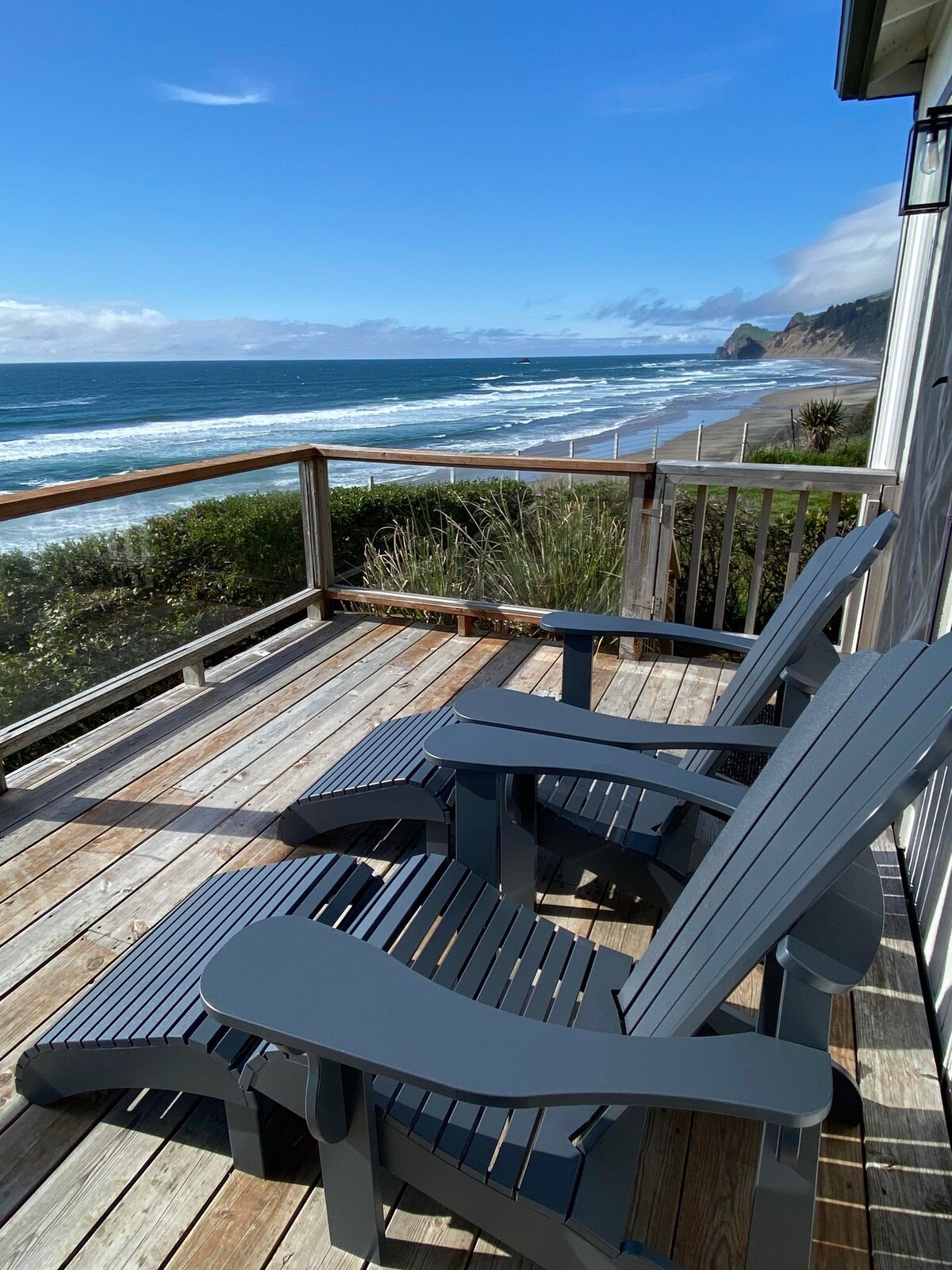 A wooden deck is furnished with two dark grey lounge chairs, offering a clear view of the expansive ocean and sandy beach below. Gentle waves roll towards the shore under a bright blue sky with scattered clouds.