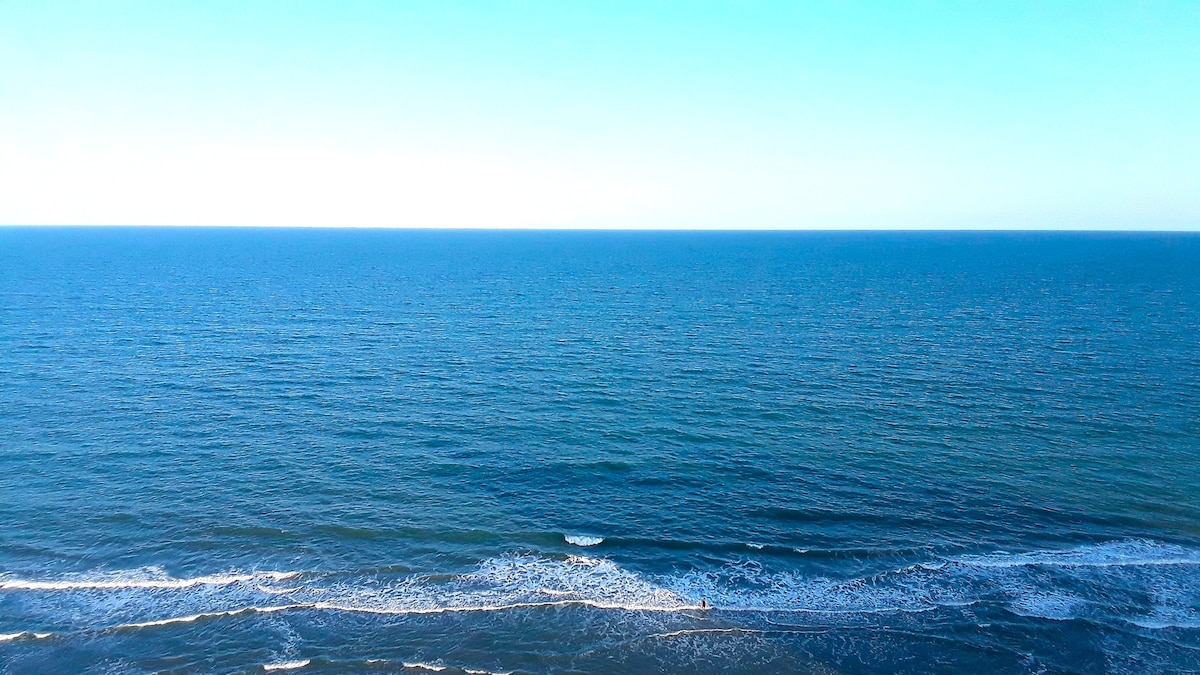 A panoramic view of the Atlantic Ocean displays a vast expanse of deep blue water, gently meeting the sandy shore. Soft waves create a subtle white foam line along the beach, under a clear sky that transitions from light to deep blue.