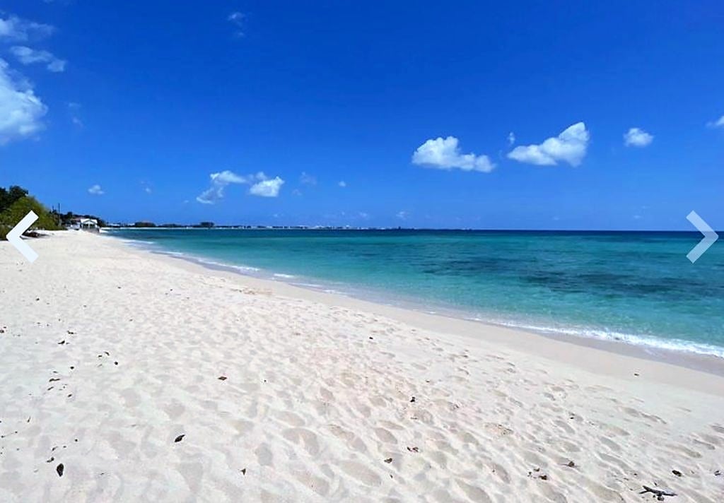 A tranquil beach scene features soft, white sand extending along the shoreline, framed by gentle waves of clear turquoise water. A bright blue sky with scattered clouds overhead completes the serene setting, inviting relaxation and enjoyment.