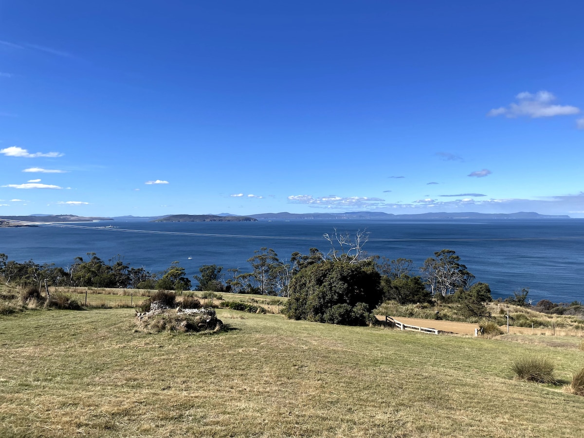 Wide panoramic views encompass the tranquil waters of Storm Bay, set against a backdrop of distant hills and a clear blue sky. The foreground features a grassy area and scattered trees, providing a serene natural setting.