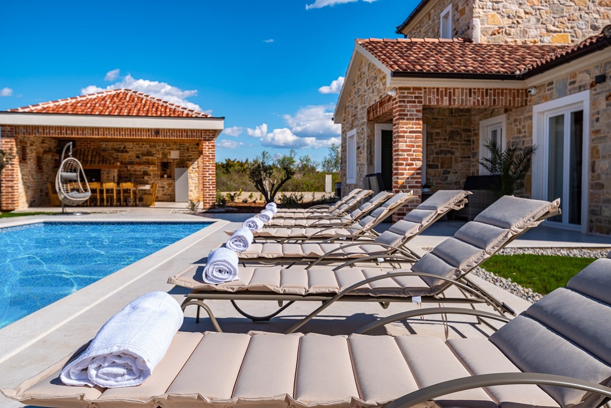 A sparkling pool is framed by several sun loungers, each topped with a neatly rolled towel. A scenic outdoor seating area with a wooden table and a covered lounge space in the background completes the inviting outdoor scene under a clear blue sky.