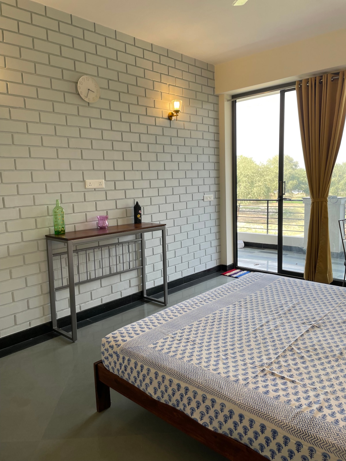 A well-lit room is presented with a double bed dressed in a patterned white and blue mattress. A wooden table stands against the wall, adorned with a green bottle and a purple glass. Large glass doors open to a balcony, offering a view of greenery.