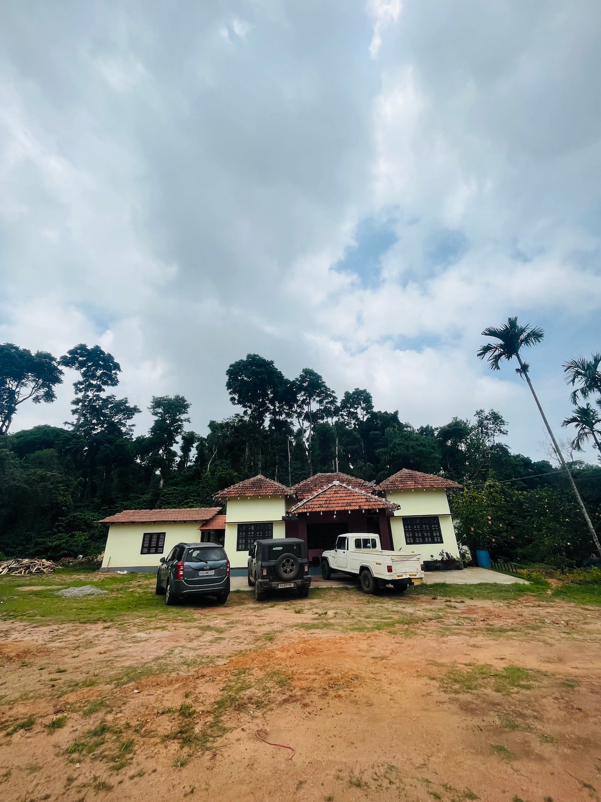 The quaint house is surrounded by lush greenery, with tall trees providing a natural backdrop. The exterior features a traditional tiled roof and multiple windows. Two vehicles are parked in front, with a gravel driveway leading to the entrance.