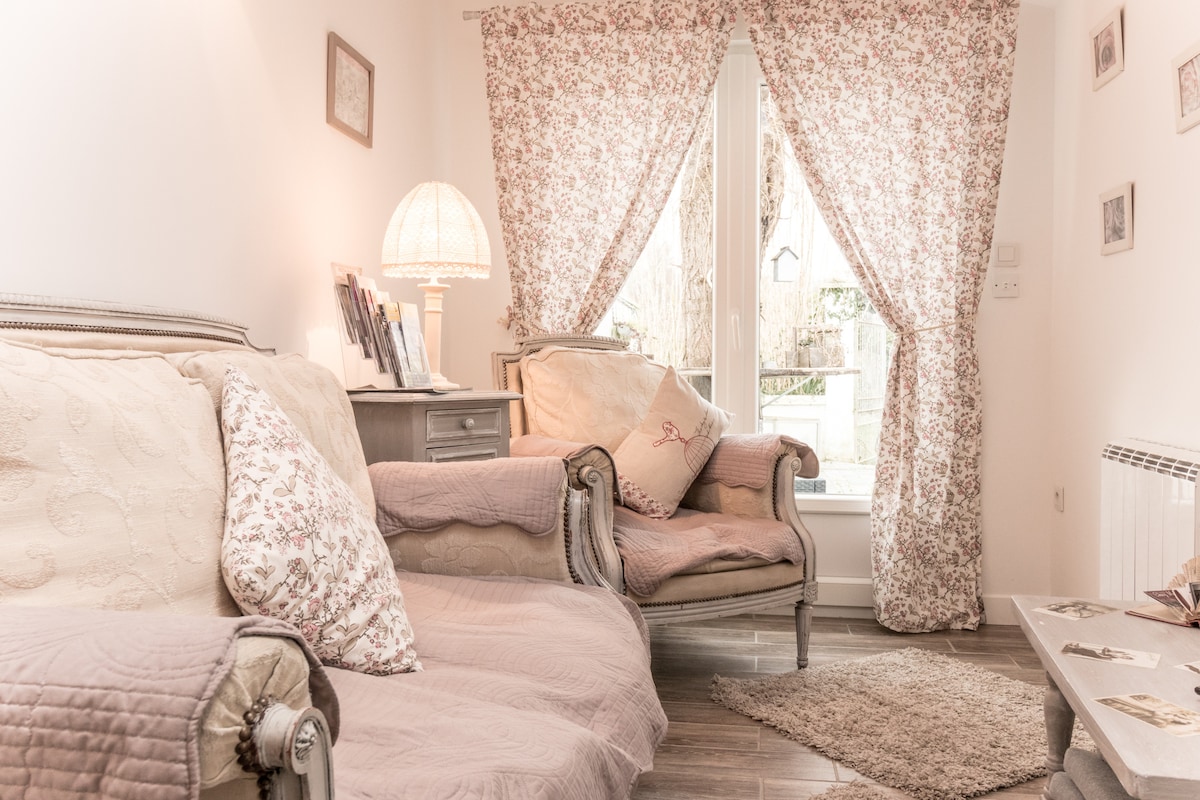 A cozy sitting area features soft seating with floral-patterned upholstery. Natural light enters through large windows dressed with matching curtains. A small table is positioned between the chairs, and decorative items are arranged on a nearby shelf, creating a welcoming environment.
