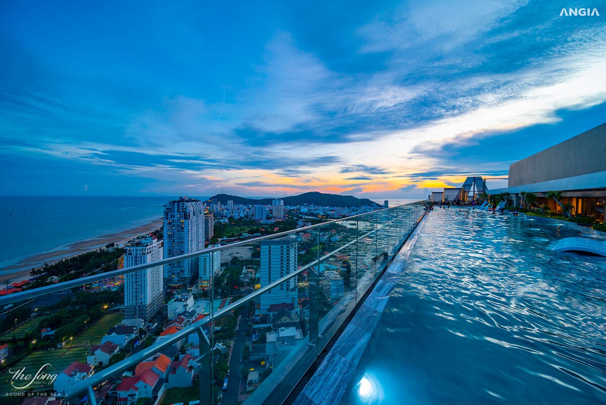 An infinity pool is situated on a rooftop, offering expansive views of the coastline and city skyline. The setting sun casts a warm glow over the horizon, enhancing the tranquil atmosphere. Lush green hills and urban landscapes create a picturesque backdrop.