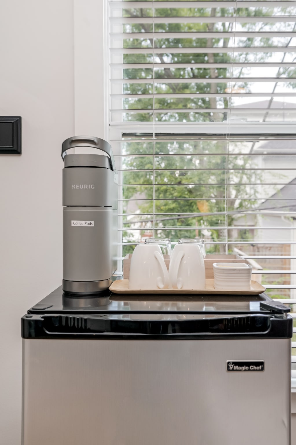 A small refrigerator is positioned beneath a countertop, topped with a Keurig coffee maker. Several white cups and saucers are arranged neatly on a tray beside the coffee machine. Natural light filters through the blinds of a nearby window, enhancing the functional setup.