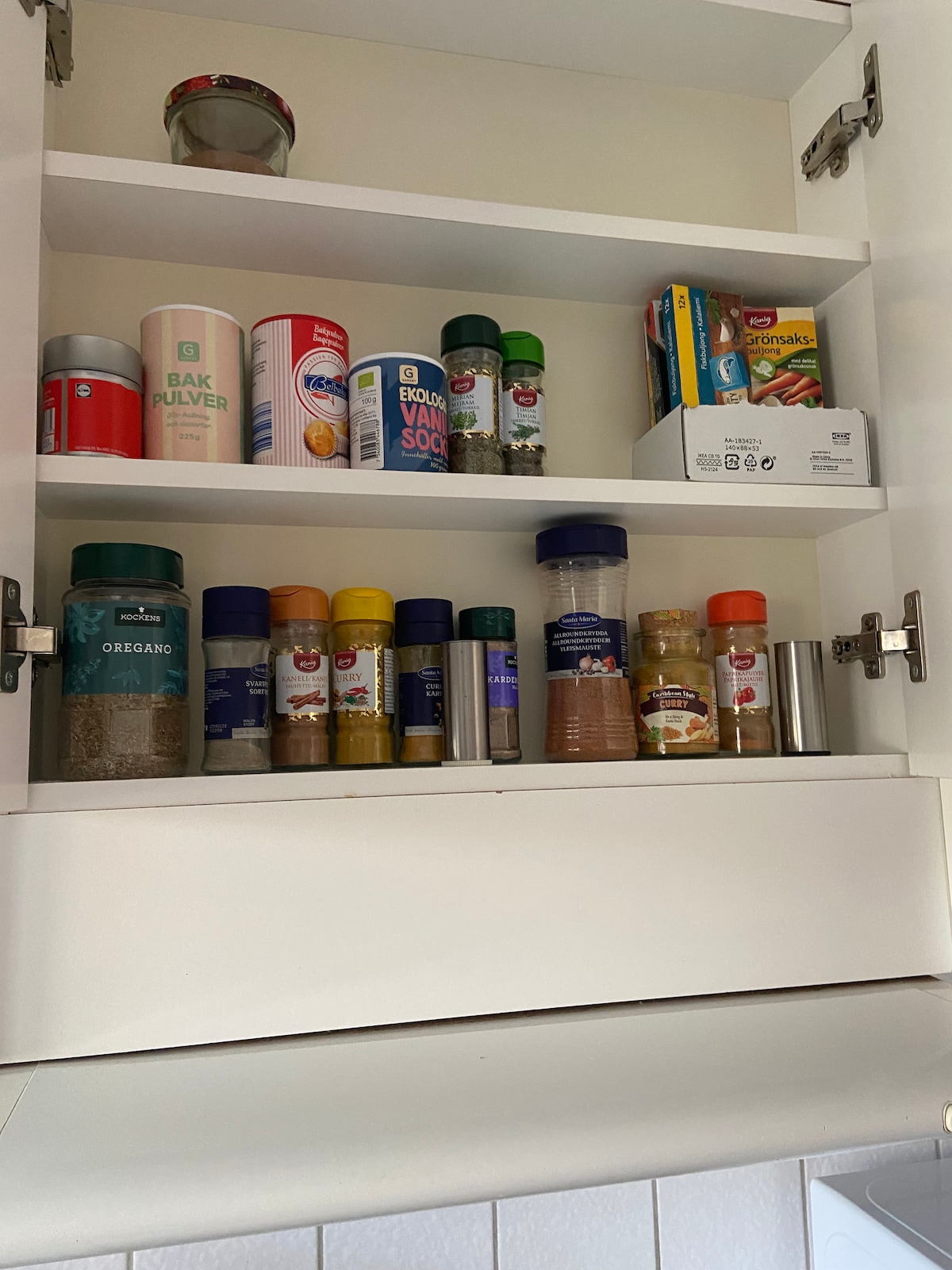 A kitchen cupboard is displayed, featuring a variety of spice containers arranged by height. Several brightly colored jars and bottles, including spices and baking essentials, are visible. A shelf above holds a few food items in boxes, completing the organized kitchen storage.