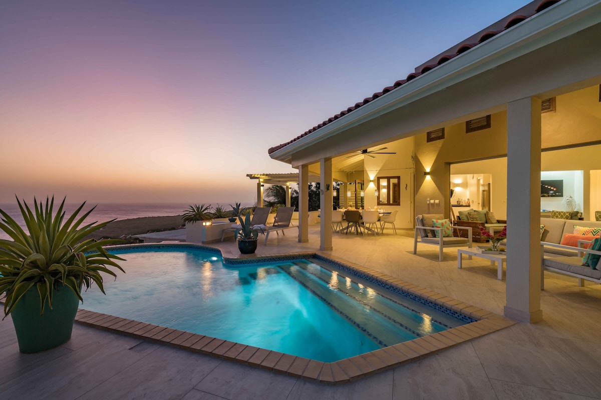 A serene view of the villa is presented at dusk, featuring a private swimming pool reflecting the soft glow of the sky. Comfortable seating areas are visible under a covered terrace, with gentle coastal breezes enhancing the peaceful atmosphere.