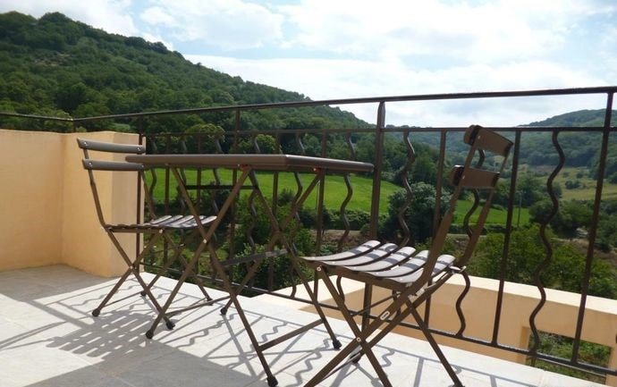 A balcony space is depicted, featuring a small table and two folding chairs, positioned against a wrought-iron railing. Lush green hills and a clear blue sky serve as the backdrop, creating an inviting area for outdoor relaxation.
