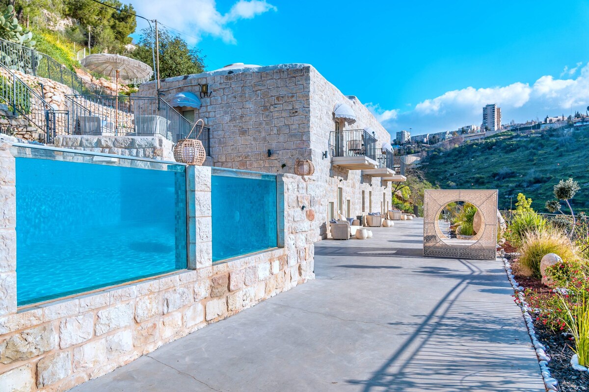 An outdoor area features a large, inviting pool with transparent walls, providing a view of the surroundings. The stone structure is complemented by well-maintained landscaping, including greenery and decorative elements. Sun loungers and umbrellas are positioned for relaxation under the open sky.