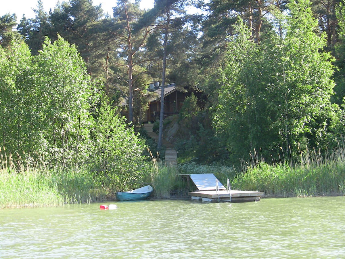 A serene lakeside view showcases a small dock and a canoe resting on calm waters. Lush greenery surrounds the area, with tall trees framing the distant cottage, creating a natural setting that emphasizes tranquility and connection with nature.