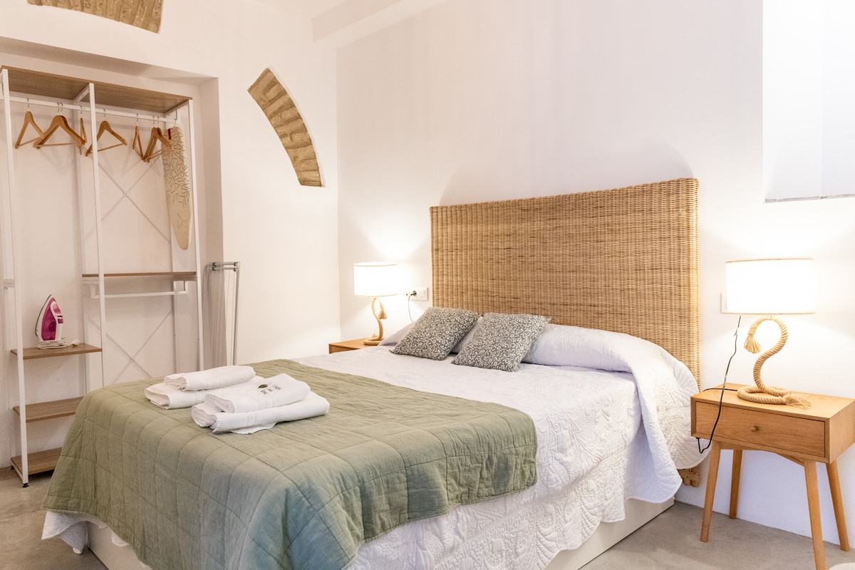 A spacious bedroom features a large bed with a textured headboard and crisp white linens. Two bedside tables hold lamps providing soft lighting. An open closet with hanging space is visible on one side, with a dressing area that includes an iron.