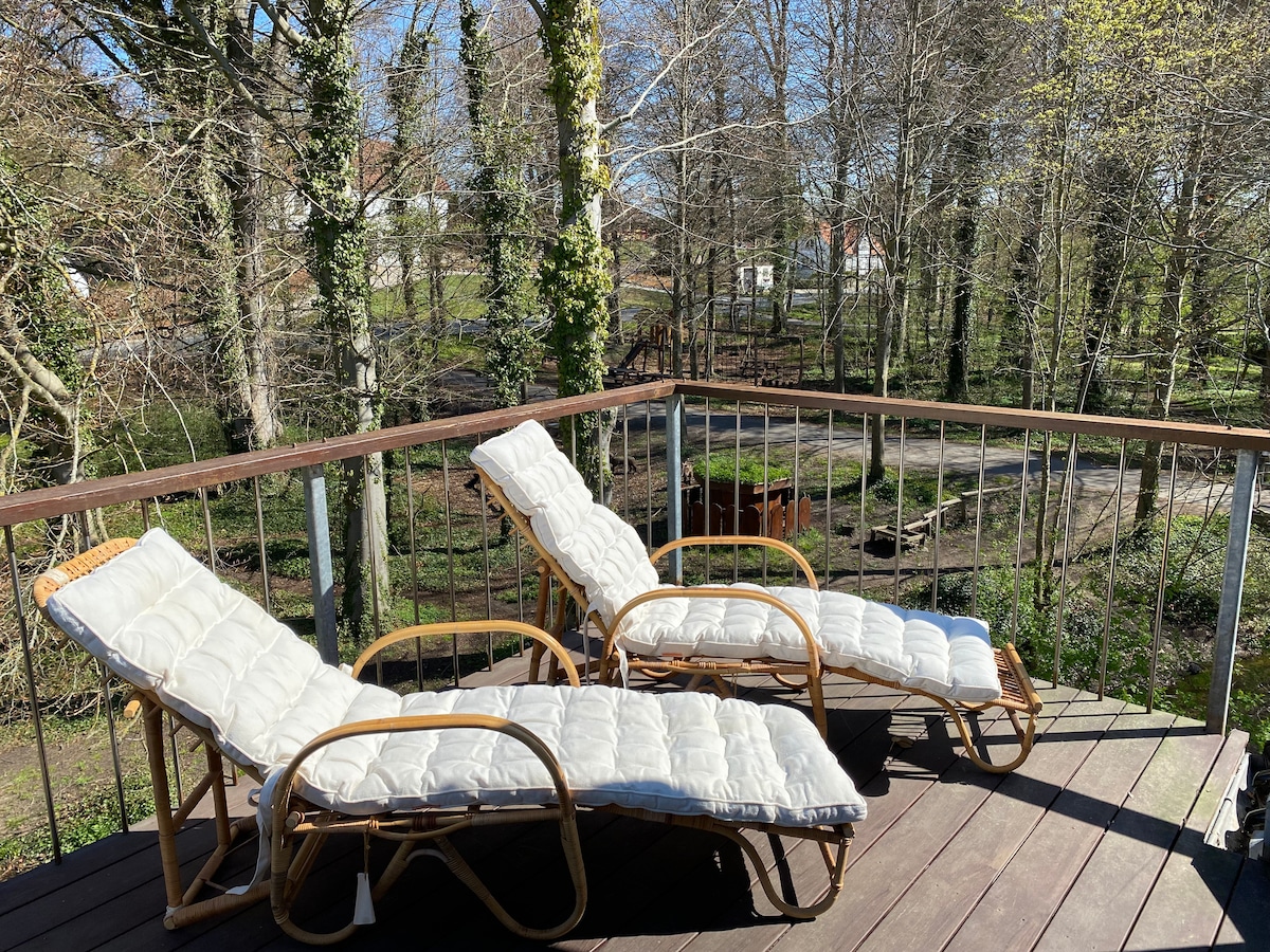Two lounge chairs are positioned on a wooden deck, offering a comfortable spot to relax. Surrounding greenery includes trees and pathways, enhancing an outdoor setting in a tranquil environment.
